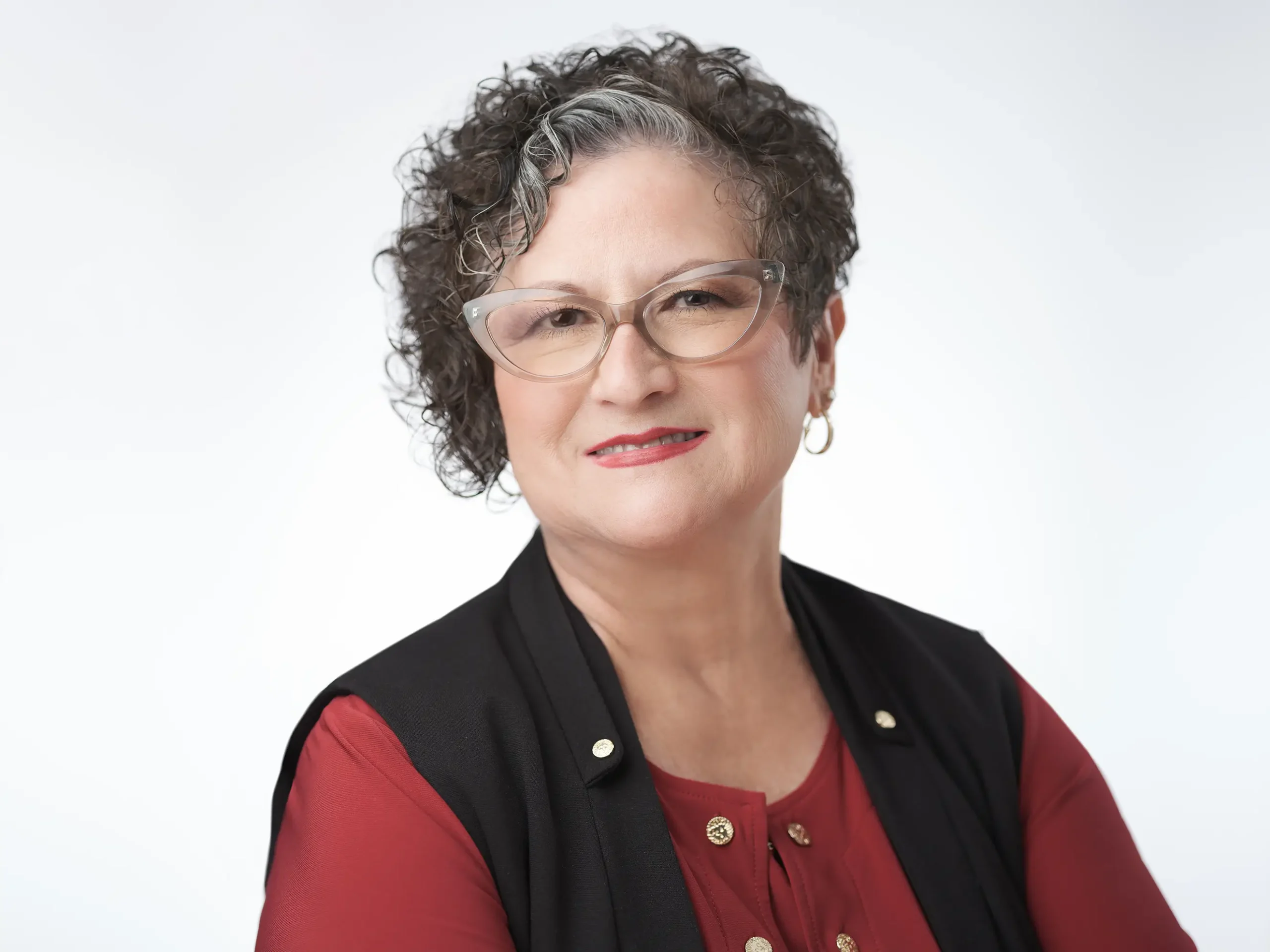 A woman with short, curly hair and wearing glasses smiles at the camera. She is dressed in a red blouse with a black vest, and gold earrings and buttons. The background is a plain, light color.