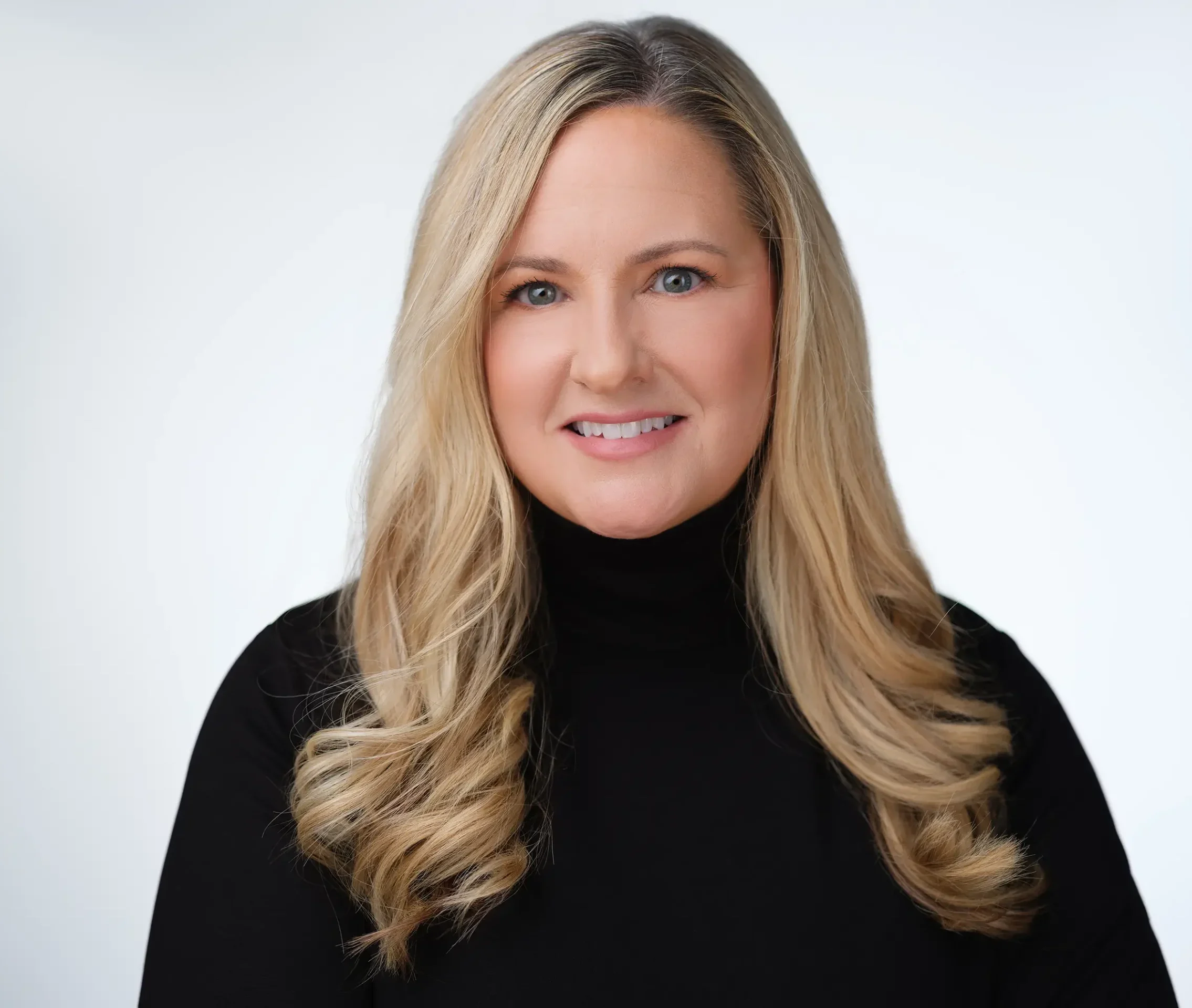 A woman with long blonde hair wearing a black turtleneck smiles at the camera against a plain light background.