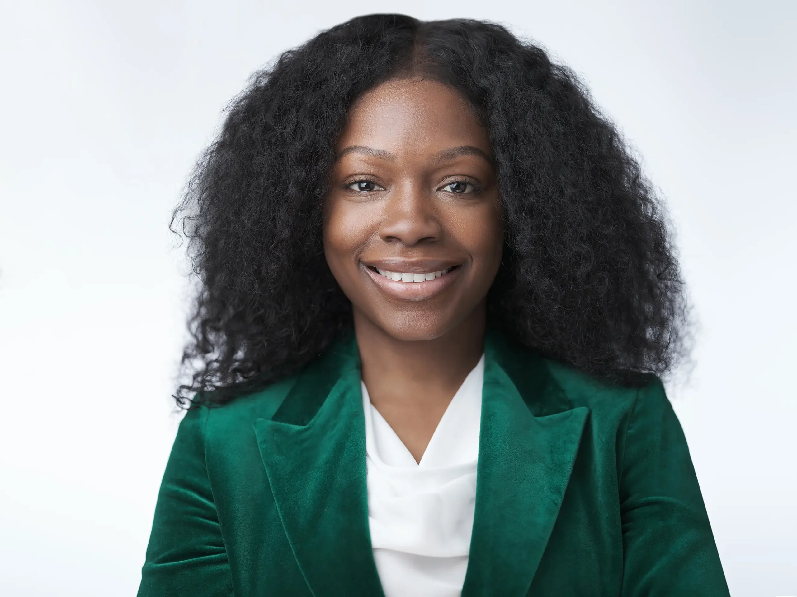 A person with long, curly hair smiles warmly, wearing a green velvet blazer over a white blouse. The background is a soft, light gray, creating a professional and approachable appearance.