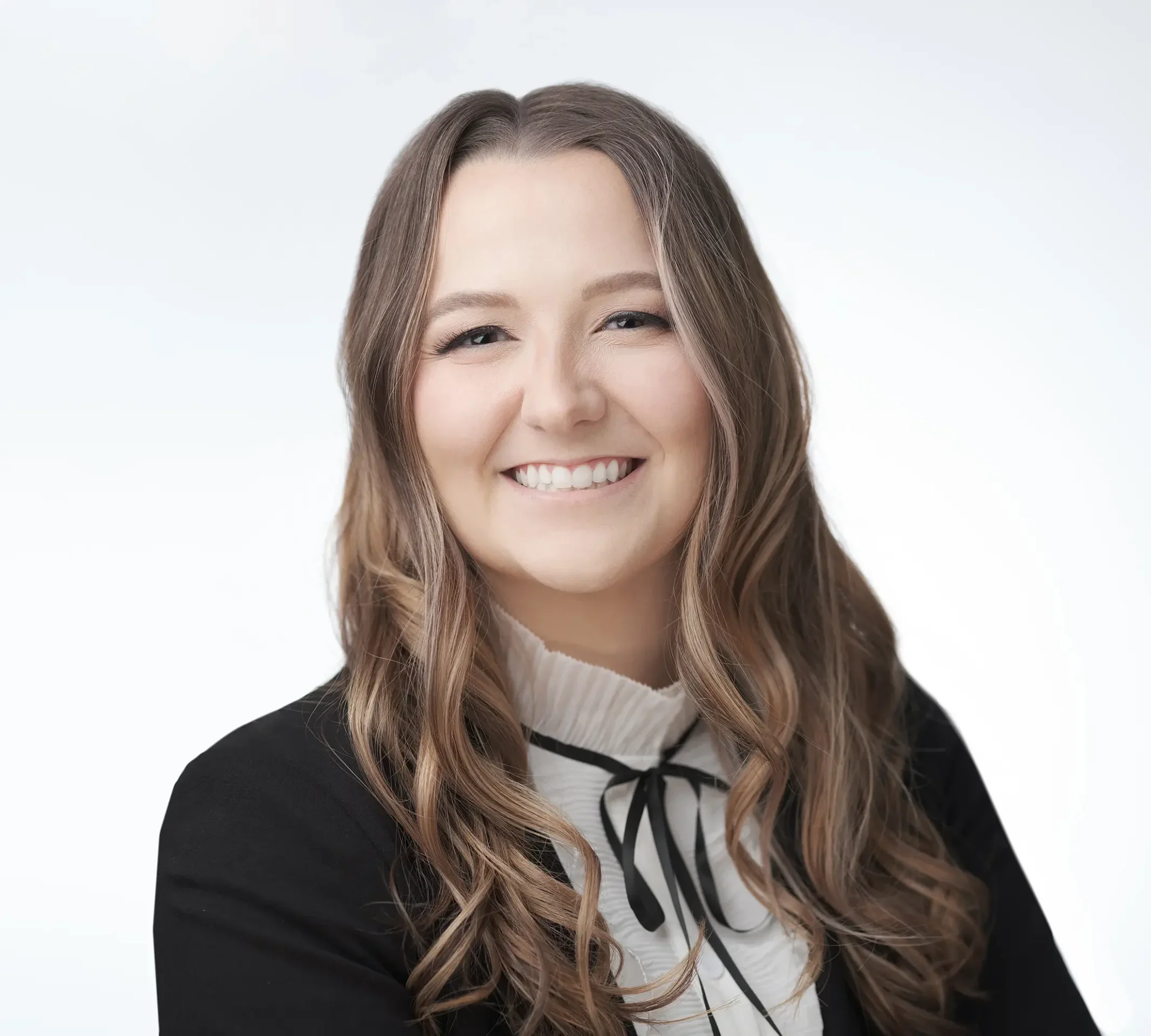 A woman with long, wavy brown hair smiles while wearing a black blazer over a white blouse with a black bow. The background is a light gray.