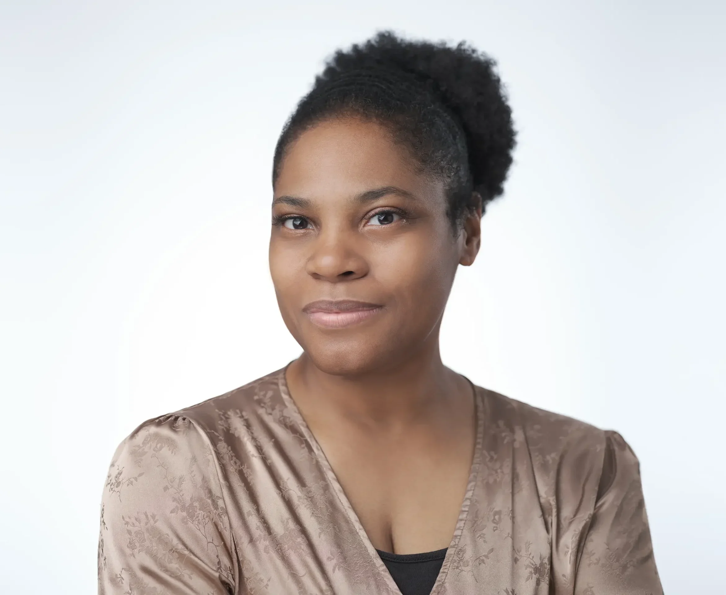 A woman with a serene expression and natural hair pulled back into a puff, wearing a light brown patterned blouse, poses against a plain white background.