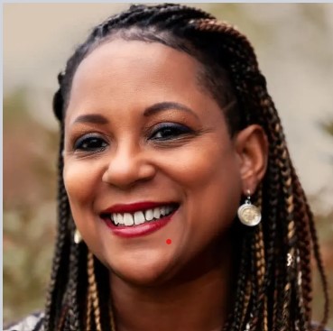 A person with braided hair smiles warmly at the camera, wearing circular earrings and red lipstick. The background is softly blurred, focusing attention on the person's cheerful expression.