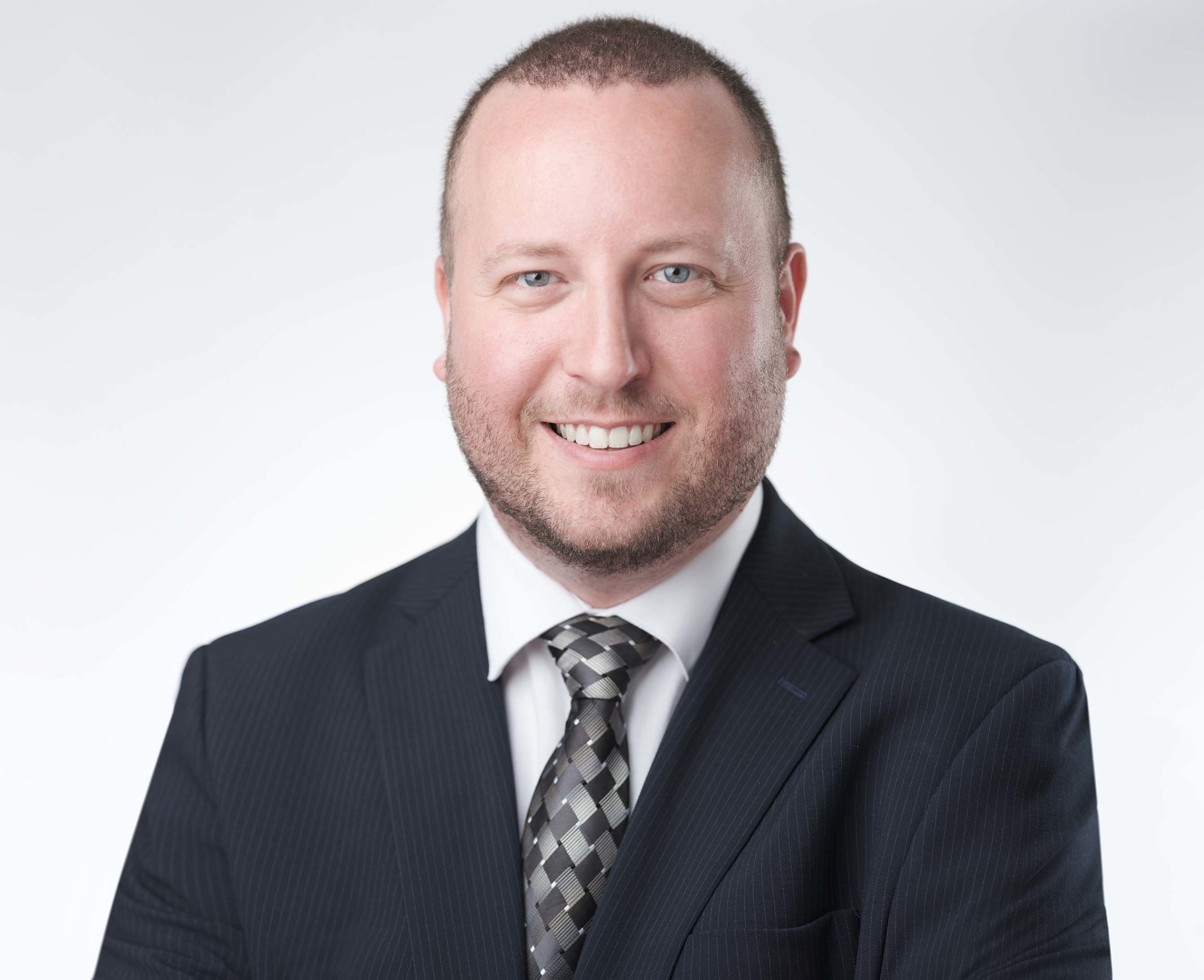 A smiling man wearing a dark suit and patterned tie stands against a plain light background. He has short hair and a trimmed beard.