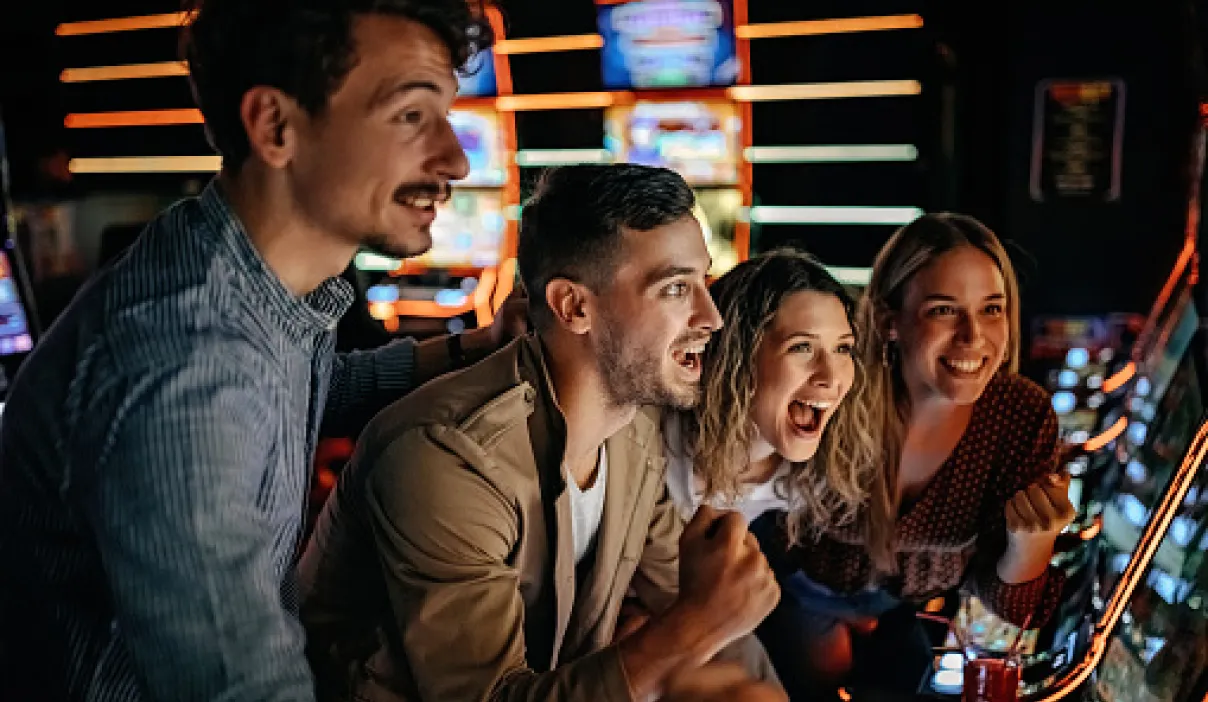 Four people excitedly play on slot machines in a dimly lit arcade. Neon lights illuminate their expressions of joy and anticipation. They are closely gathered, focusing intently on the machines.