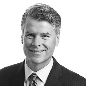 A black and white portrait of a man in a suit and tie, smiling. He has short, neatly styled hair and is looking directly at the camera. The background is plain white.