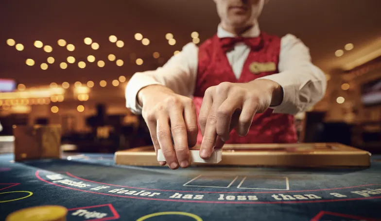 Casino dealer in a red vest shuffling cards at a blackjack table. Blurred background with bokeh lights enhances the casino ambiance.