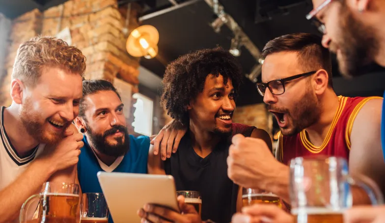 A group of five men sitting at a bar table, enjoying drinks and laughing. One is holding a tablet, while another excitedly reacts. The setting has a warm, rustic ambiance with exposed brick walls and hanging lights.