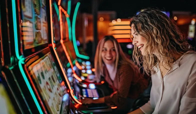 Two people are enjoying themselves at a casino. They are seated side by side at slot machines, surrounded by vibrant neon lights. Both appear engaged and happy, with the atmosphere lively and colorful.