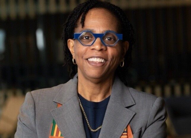 Person with short braided hair wearing blue glasses, a gray blazer with colorful patterned accents, a dark shirt, and a gold necklace. They are smiling with arms crossed. The background is a blurred indoor setting.