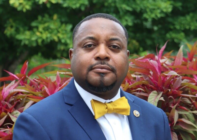 Man in a blue suit and yellow bow tie sits in front of deep red foliage with green trees in the background. He has a neatly trimmed beard and a white shirt, looking directly at the camera with a calm expression.