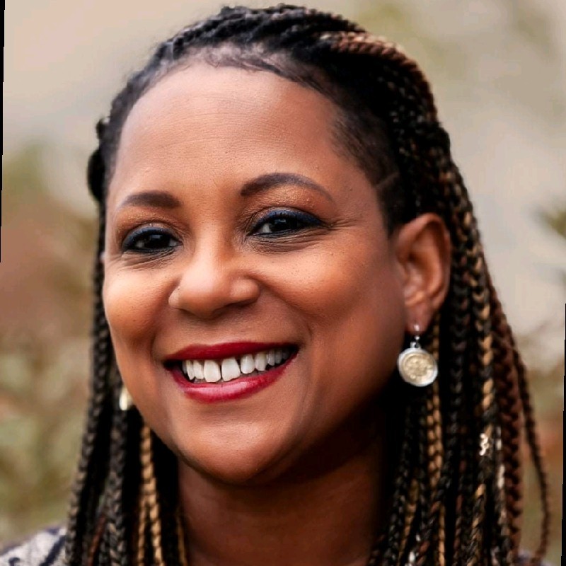Smiling woman with braided hair wearing circular earrings and a patterned top. The background is softly blurred.