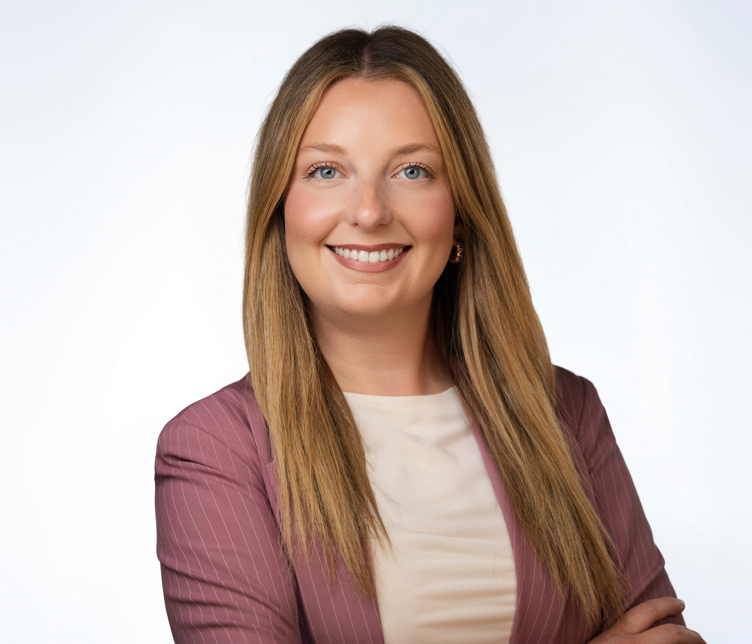 A woman with long straight blonde hair, wearing a mauve blazer and white top, stands smiling with arms crossed against a plain light background.