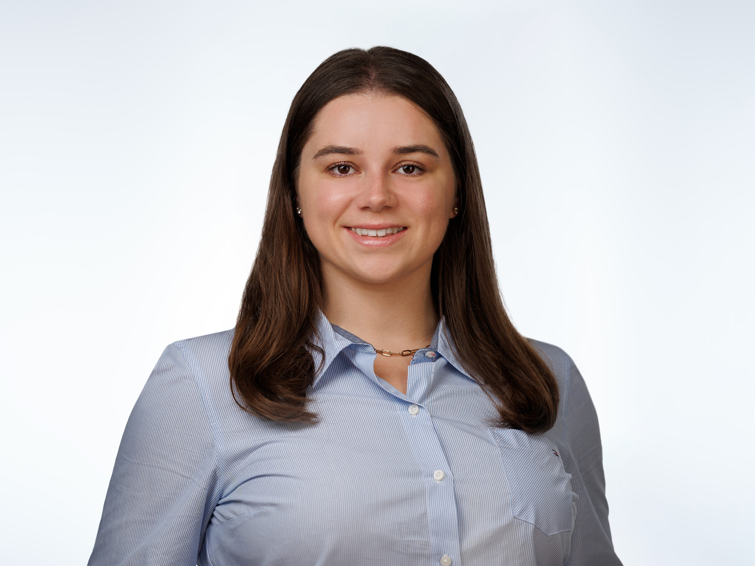 A young woman with long brown hair, wearing a light blue button-up shirt and a gold necklace, smiles at the camera against a plain, light background.