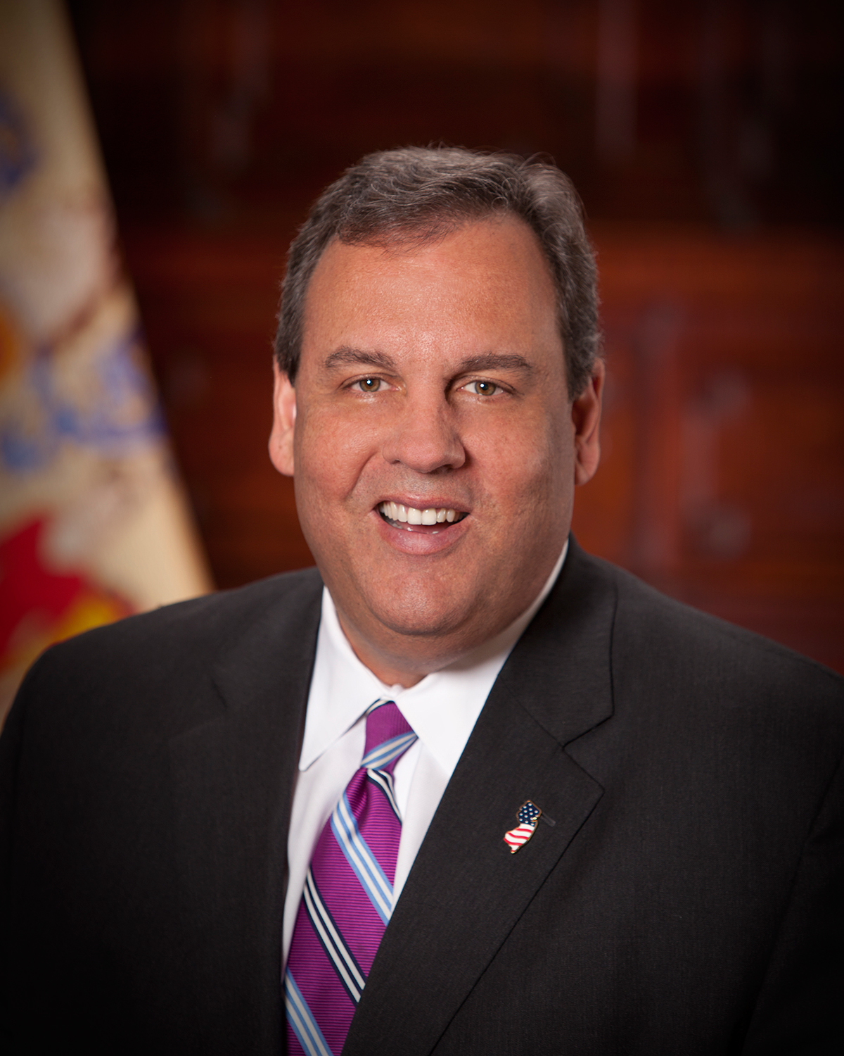 A man in a dark suit, white shirt, and purple striped tie smiles at the camera. He wears a U.S. flag pin on his lapel, and a blurred flag is visible in the background.