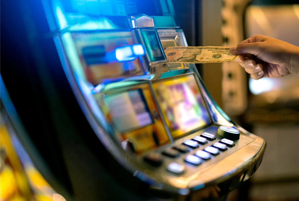 A person’s hand inserts a U.S. twenty-dollar bill into a brightly lit slot machine, with buttons and screens visible, in a casino setting.