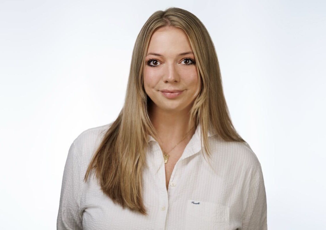 A woman with long blonde hair wearing a white collared shirt against a white background.